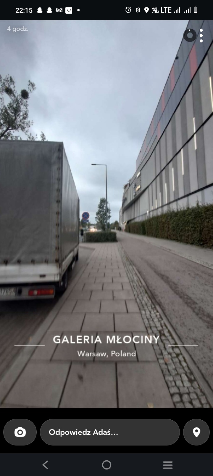 Szary bus transportowy zaparkowany na chodniku przy Galerii Młociny w Warszawie, Polska. Ujęcie z perspektywy chodnika, widoczny fragment budynku galerii.