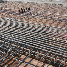 Siatka z prętów stalowych, tworząca zbrojenie płyty fundamentowej, widoczna pod kątem, z elementami dystansowymi. Przygotowanie do zalewania betonem.