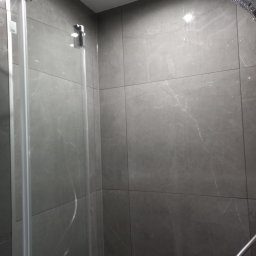 Anton Kanonchyk - Szklana kabina prysznicowa z deszczownicą i słuchawką, wykończona szarymi płytkami imitującymi marmur. Widoczne chromowane elementy armatury.