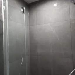 Szklana kabina prysznicowa z deszczownicą i słuchawką, wykończona szarymi płytkami imitującymi marmur. Widoczne chromowane elementy armatury.