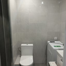 Anton Kanonchyk - Jasna łazienka po remoncie: kabina prysznicowa, toaleta i pralka na tle szarych płytek. Widoczne gniazdko elektryczne. Minimalistyczny design.