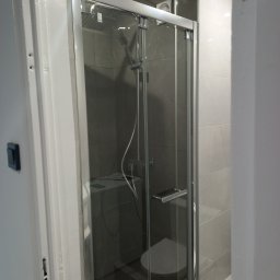 Anton Kanonchyk - Szklana kabina prysznicowa z deszczownicą w nowoczesnej łazience wykończonej szarymi płytkami. Widoczna toaleta i pralka. Minimalistyczny design.