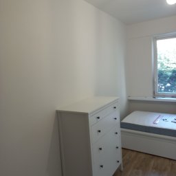 Anton Kanonchyk - Biała komoda z szufladami w pokoju z drewnianą podłogą i oknem. Ściany pomalowane na biało. Minimalistyczny styl. Widok na fragment łóżka.