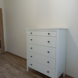 Anton Kanonchyk - Biała komoda z pięcioma szufladami i czarnymi gałkami, ustawiona na panelach podłogowych w jasnym pokoju. Widoczny fragment drzwi po lewej stronie.