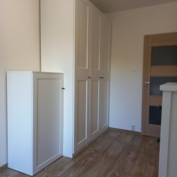 Anton Kanonchyk - Biała, wysoka szafa z czterema drzwiami i mała szafka obok, na podłodze panele imitujące drewno, w tle drzwi z szybą. Minimalistyczny styl.