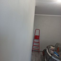 Anton Kanonchyk - Świeżo pomalowane na szaro ściany pokoju z czerwoną drabiną opartą o ścianę. Meble zabezpieczone folią malarską. Widoczne panele podłogowe.