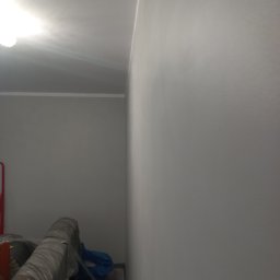 Anton Kanonchyk - Świeżo pomalowana, gładka ściana w odcieniach szarości z widoczną listwą przypodłogową i fragmentem sufitu. W tle zrolowane materiały budowlane.