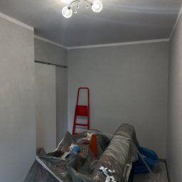 Anton Kanonchyk - Pokój po malowaniu na szaro, zabezpieczone meble folią, czerwona drabina oparta o ścianę, widoczna lampa sufitowa. Świeżo pomalowane ściany i listwy.
