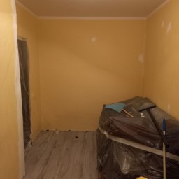 Anton Kanonchyk - Pokój w trakcie malowania na żółto, z zabezpieczonymi folią meblami i widocznym odkurzaczem. Świeżo położone panele podłogowe i pomalowane listwy przypodłogowe.