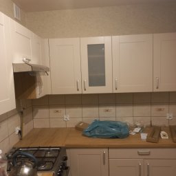 Anton Kanonchyk - Jasna kuchnia z beżowymi szafkami i drewnianym blatem. Na ścianie płytki i tapeta. Widoczne akcesoria kuchenne i elementy montażowe. Ujęcie wnętrza po częściowym remoncie.