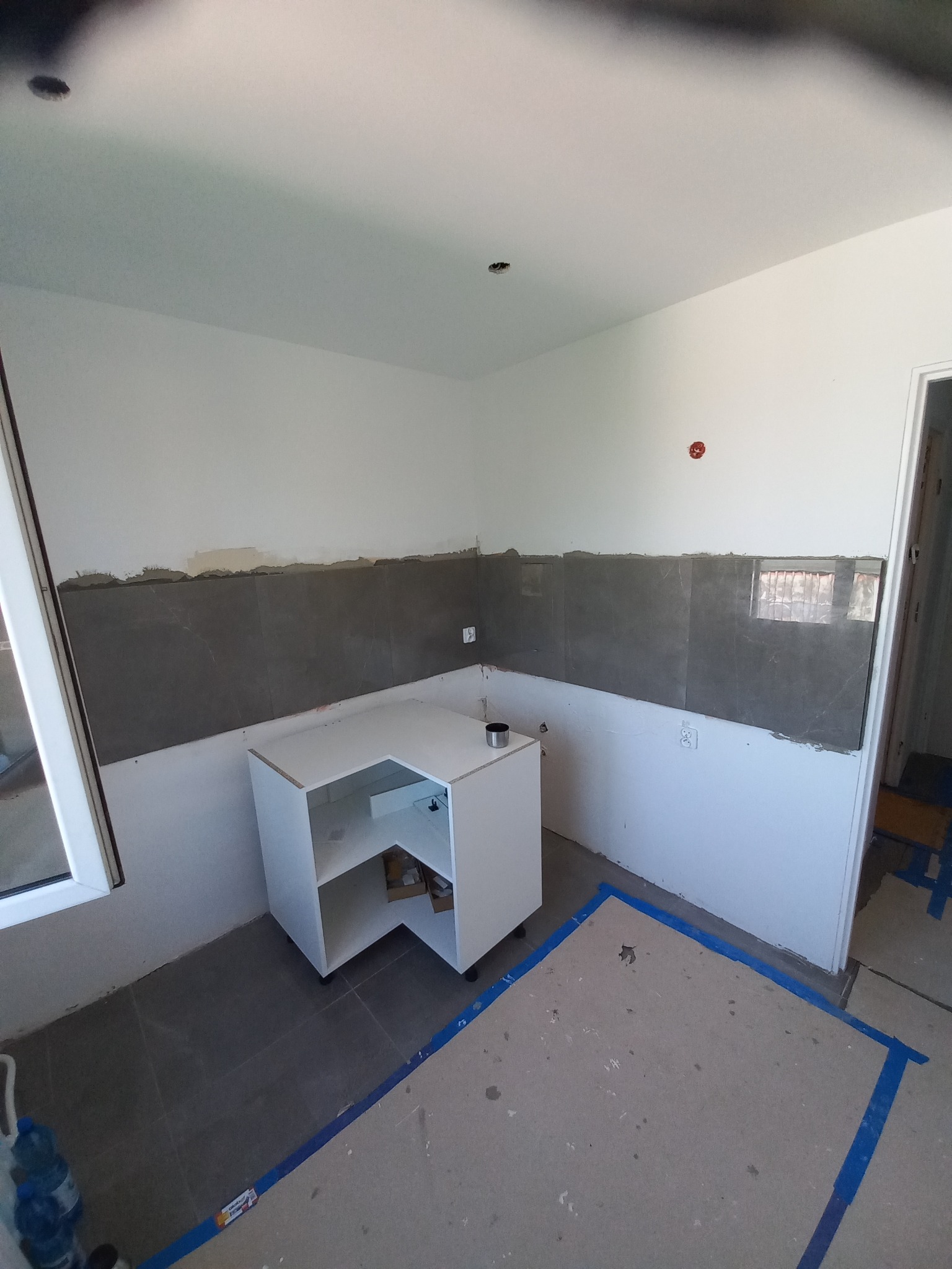 Wnętrze w trakcie prac: częściowo ułożone szare płytki na ścianie, biała szafka narożna bez frontów, zabezpieczona podłoga. Widoczne ślady prac wykończeniowych.