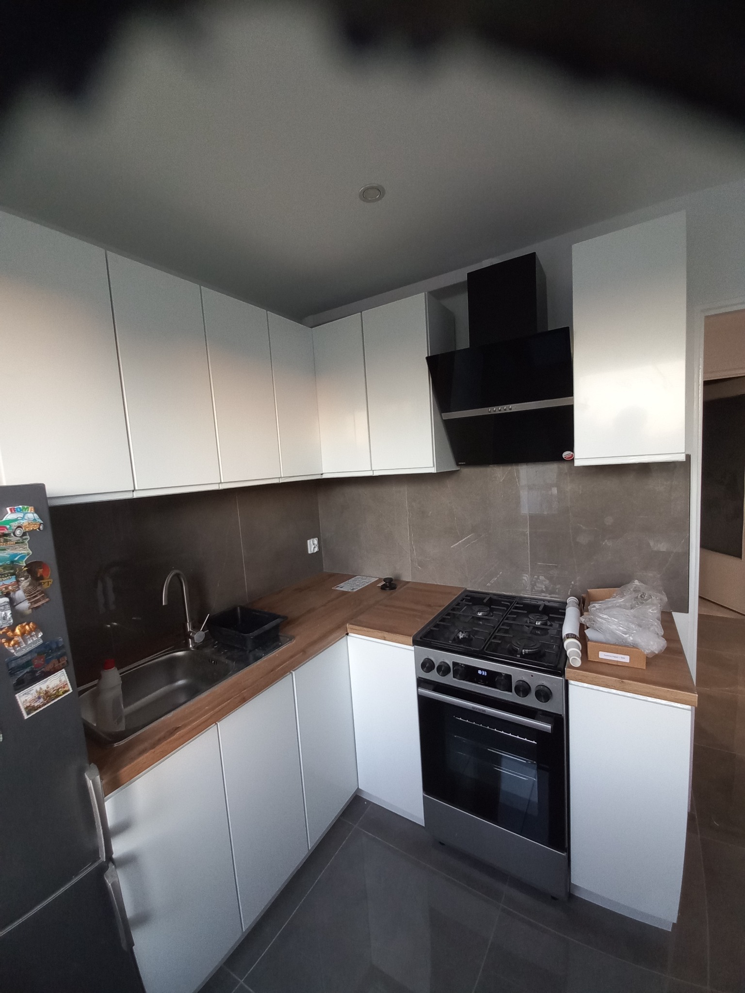 Nowoczesna kuchnia po montażu: białe szafki z drewnianym blatem, szara glazura, czarny okap nad kuchenką. Widoczne akcesoria montażowe.