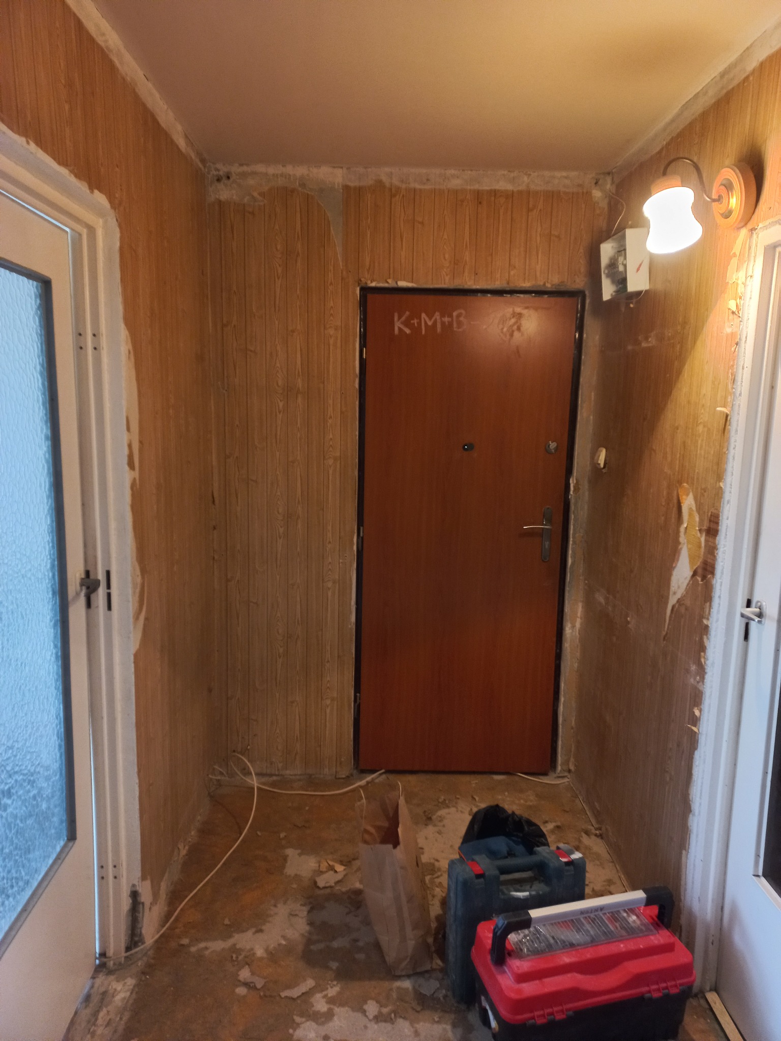 Korytarz w trakcie remontu: ściany z boazerią częściowo zerwaną, widoczne narzędzia i kable na podłodze. Na drzwiach napisane kredą 'K+M+B+'.