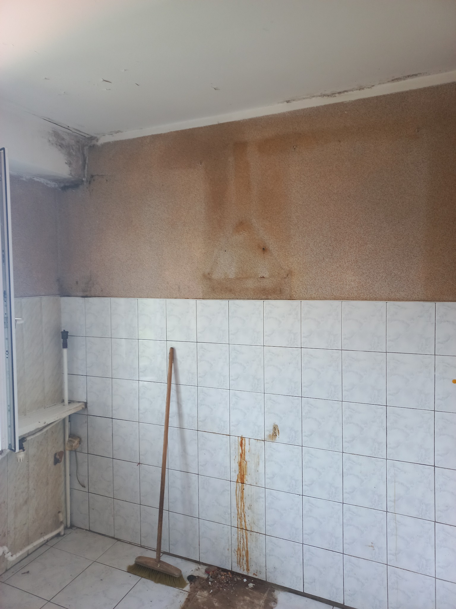 Zniszczona łazienka przed remontem: białe płytki z zaciekami, brudna podłoga, ściany z plamami i grzybem. Stara miotła oparta o ścianę. Widoczne ślady wilgoci i zaniedbania.