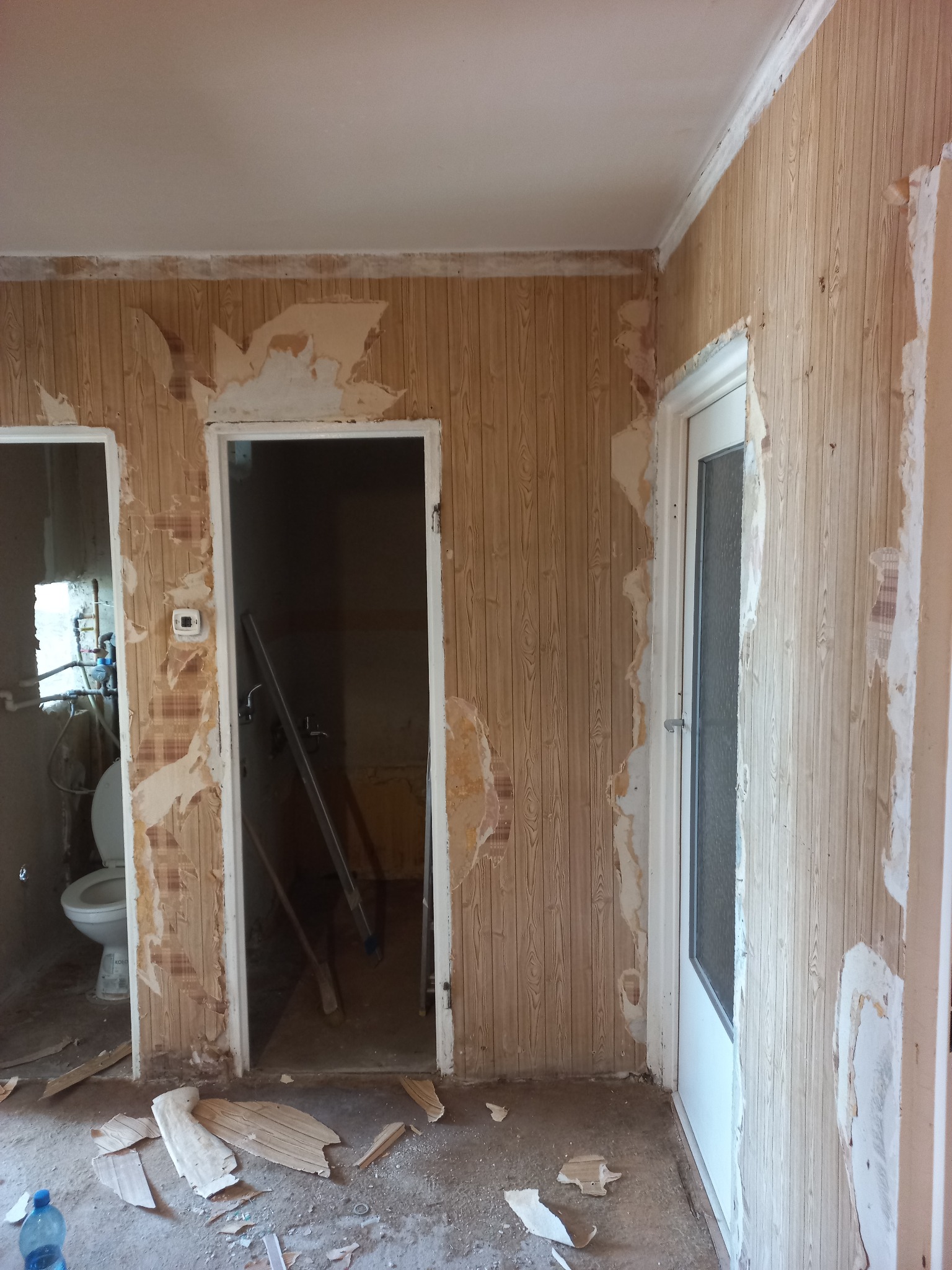 Pokój w trakcie demontażu boazerii, widoczne fragmenty oderwanych paneli na ścianach i podłodze. W tle widoczna łazienka z toaletą i drabina oparta o ścianę.