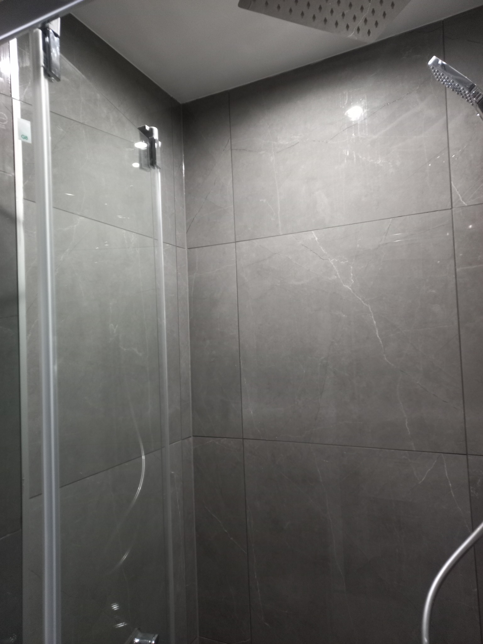 Szklana kabina prysznicowa z deszczownicą i słuchawką, wykończona szarymi płytkami imitującymi marmur. Widoczne chromowane elementy armatury.