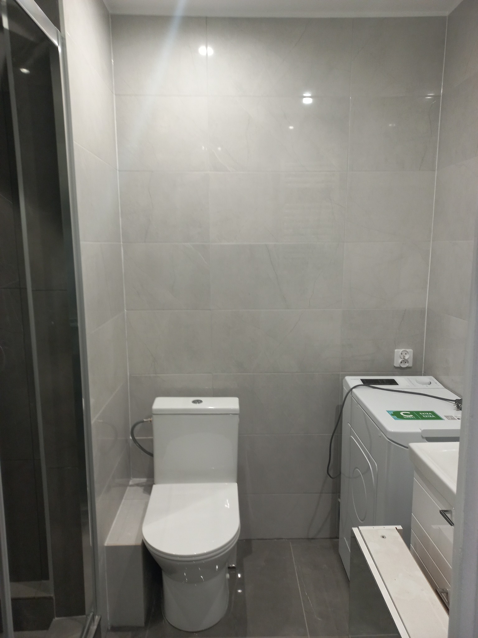 Jasna łazienka po remoncie: kabina prysznicowa, toaleta i pralka na tle szarych płytek. Widoczne gniazdko elektryczne. Minimalistyczny design.