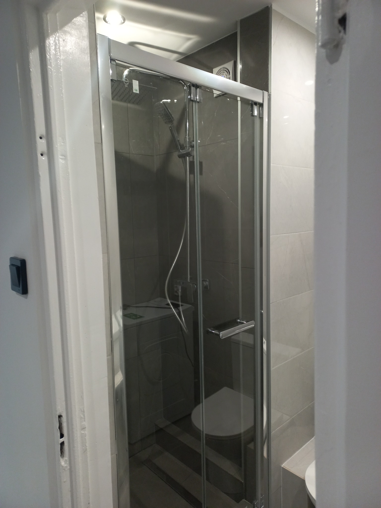 Szklana kabina prysznicowa z deszczownicą w nowoczesnej łazience wykończonej szarymi płytkami. Widoczna toaleta i pralka. Minimalistyczny design.