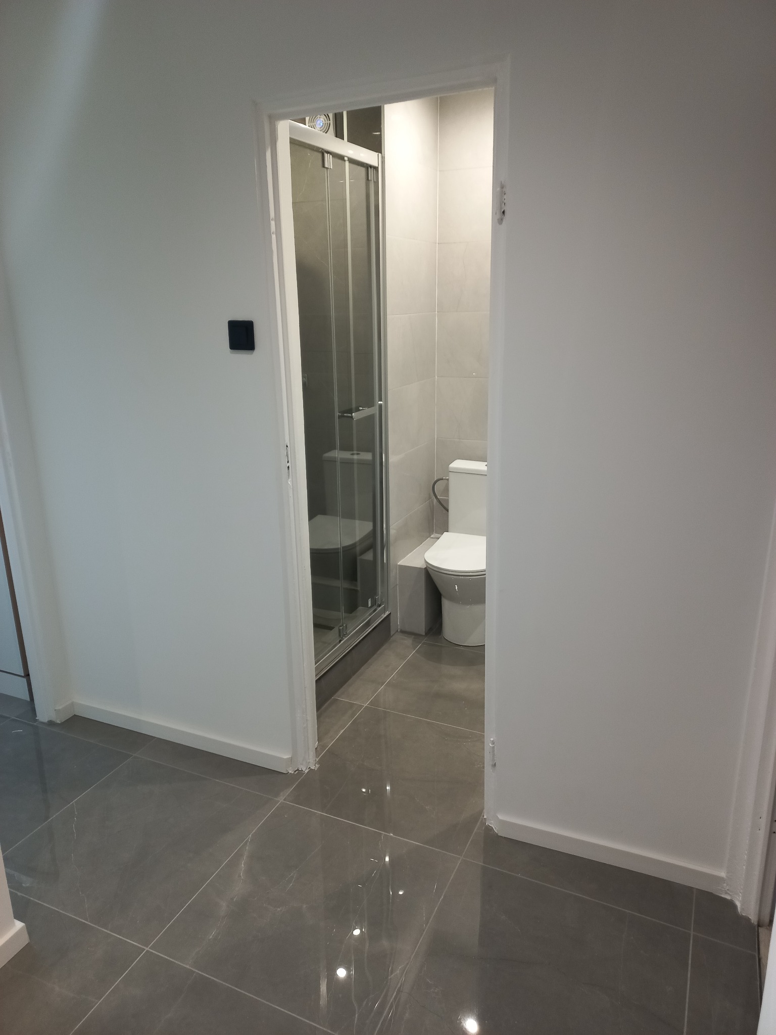 Nowoczesna łazienka po remoncie: szare płytki podłogowe, białe ściany, kabina prysznicowa z przesuwnymi drzwiami i toaleta. Minimalistyczny design, czyste linie.