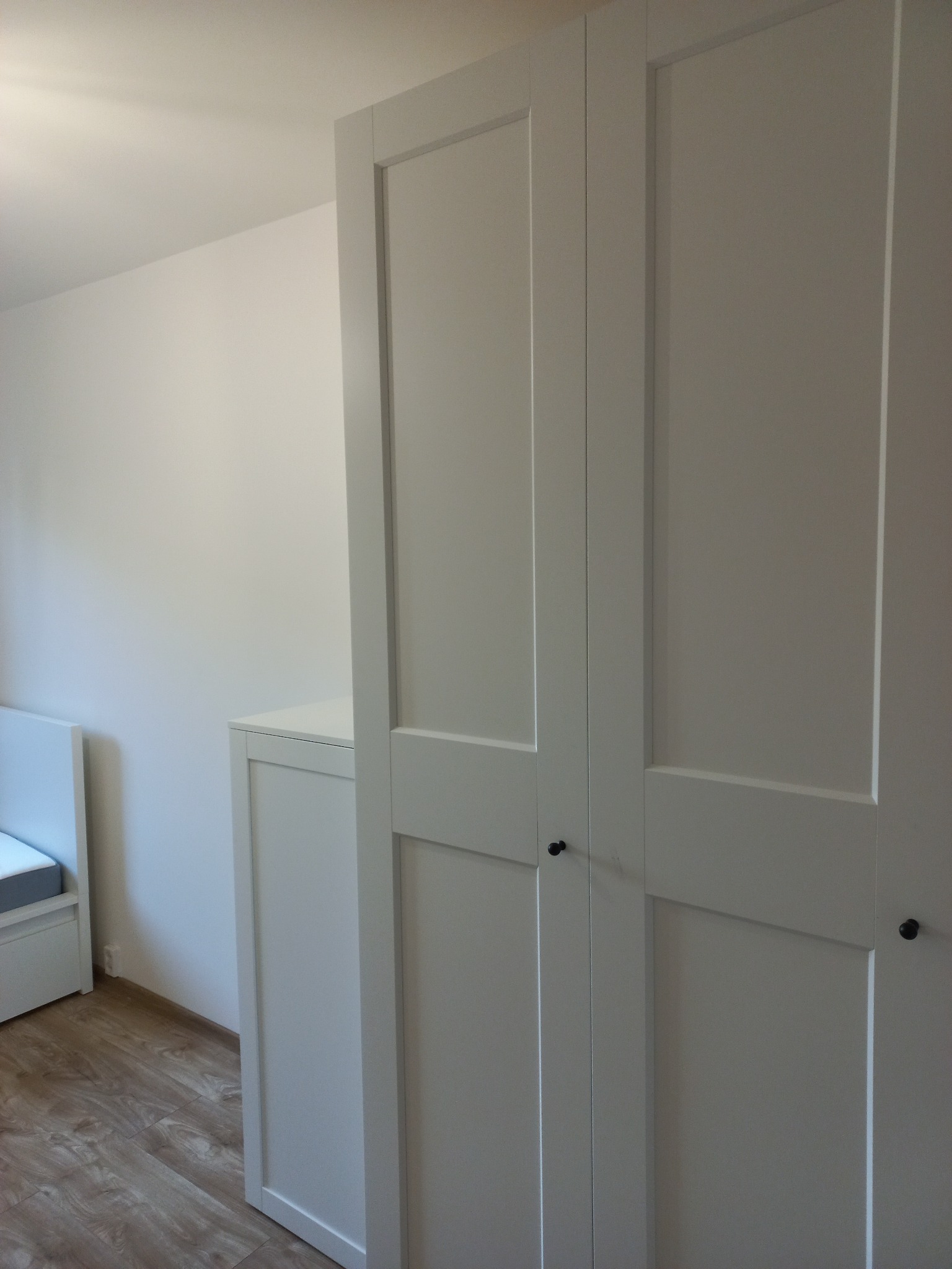 Biała, trzydrzwiowa szafa z czarnymi gałkami, wkomponowana w jasne wnętrze. Widoczny fragment łóżka i drewniana podłoga. Minimalistyczny design.