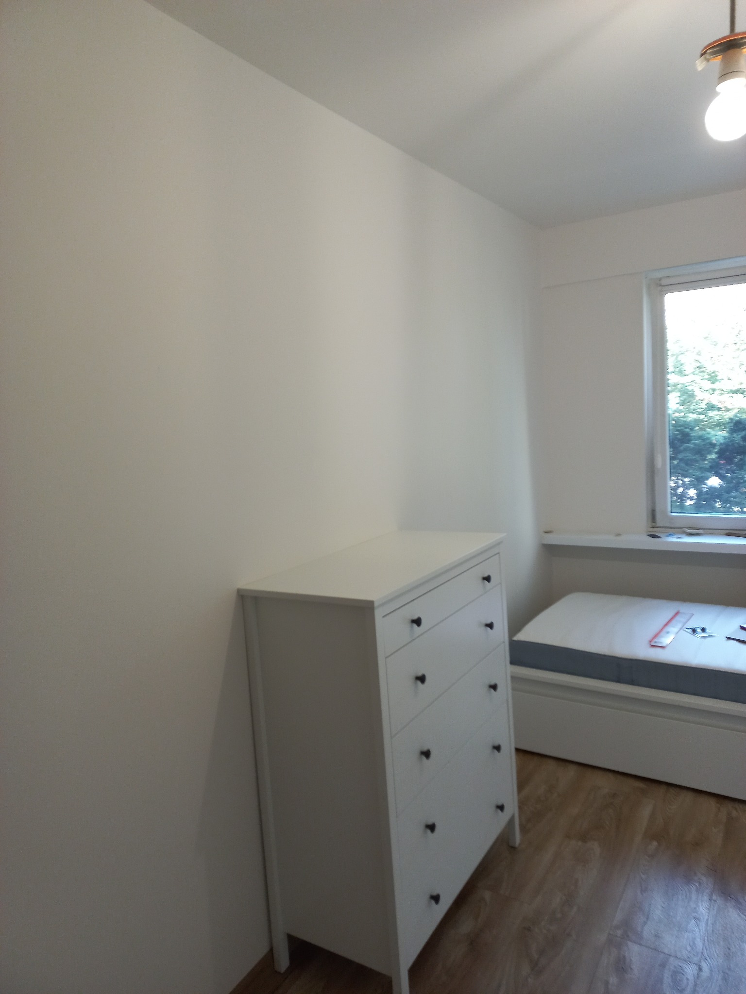 Biała komoda z szufladami w pokoju z drewnianą podłogą i oknem. Ściany pomalowane na biało. Minimalistyczny styl. Widok na fragment łóżka.