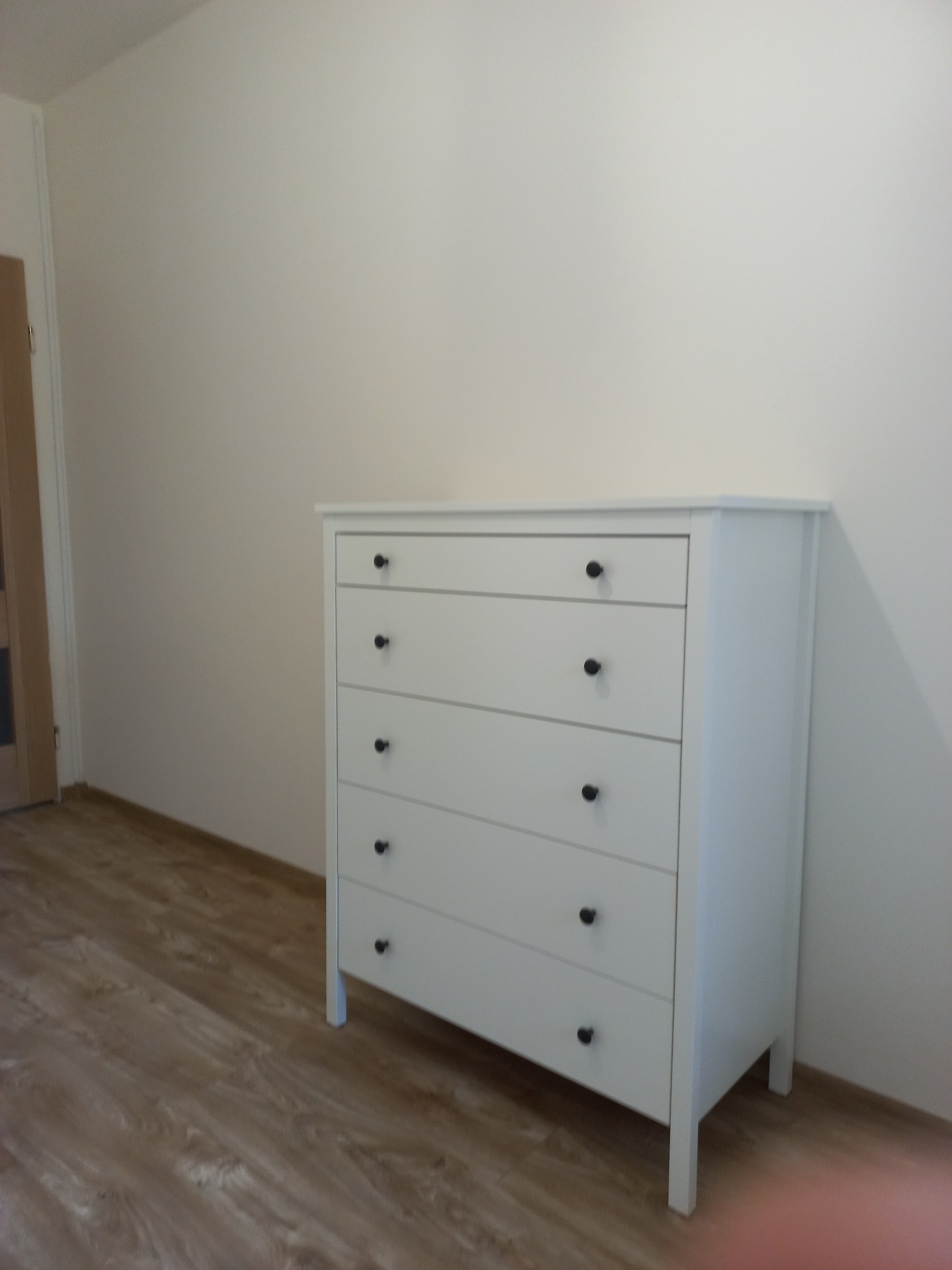 Biała komoda z pięcioma szufladami i czarnymi gałkami, ustawiona na panelach podłogowych w jasnym pokoju. Widoczny fragment drzwi po lewej stronie.