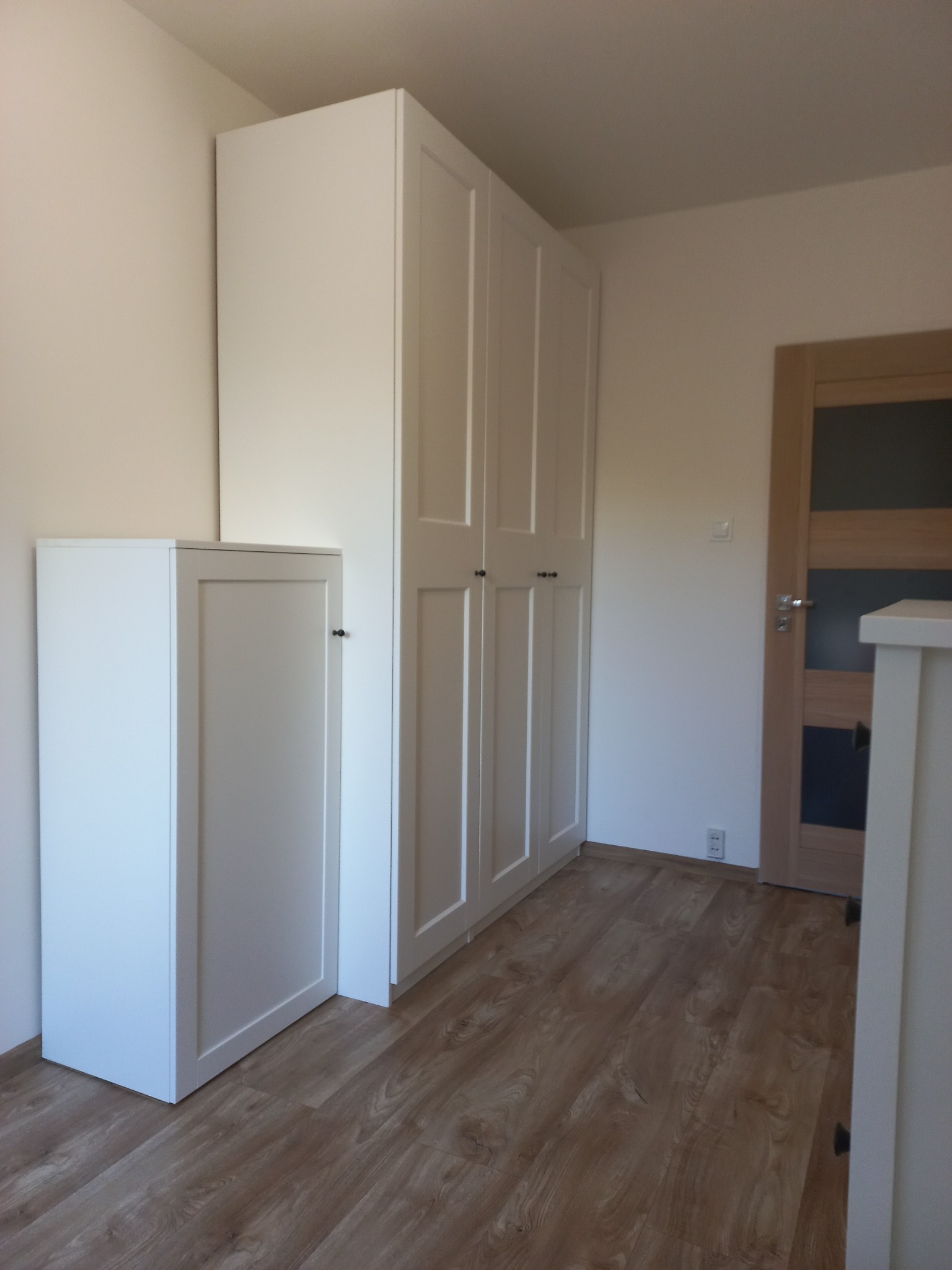 Biała, wysoka szafa z czterema drzwiami i mała szafka obok, na podłodze panele imitujące drewno, w tle drzwi z szybą. Minimalistyczny styl.