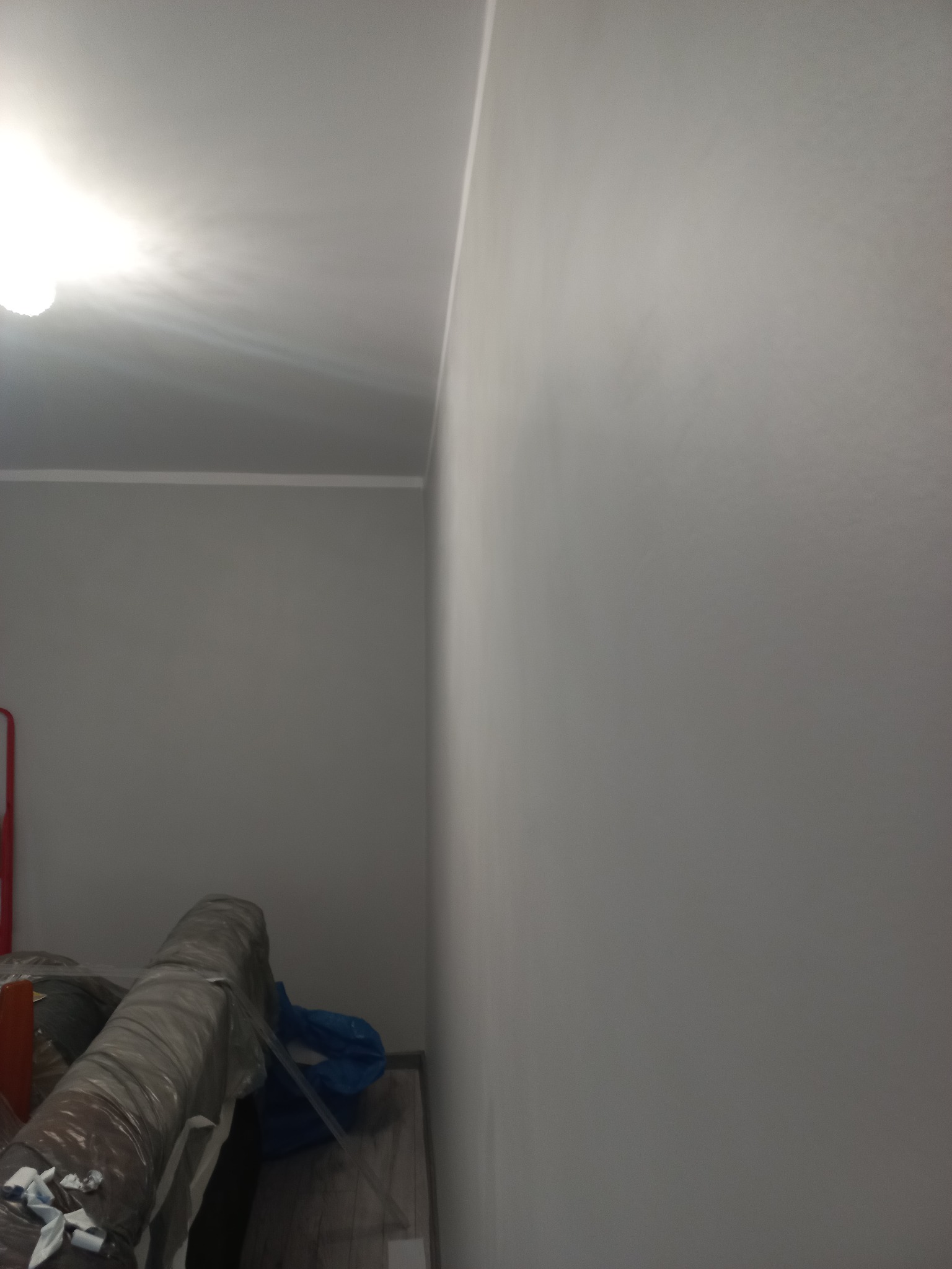 Świeżo pomalowana, gładka ściana w odcieniach szarości z widoczną listwą przypodłogową i fragmentem sufitu. W tle zrolowane materiały budowlane.