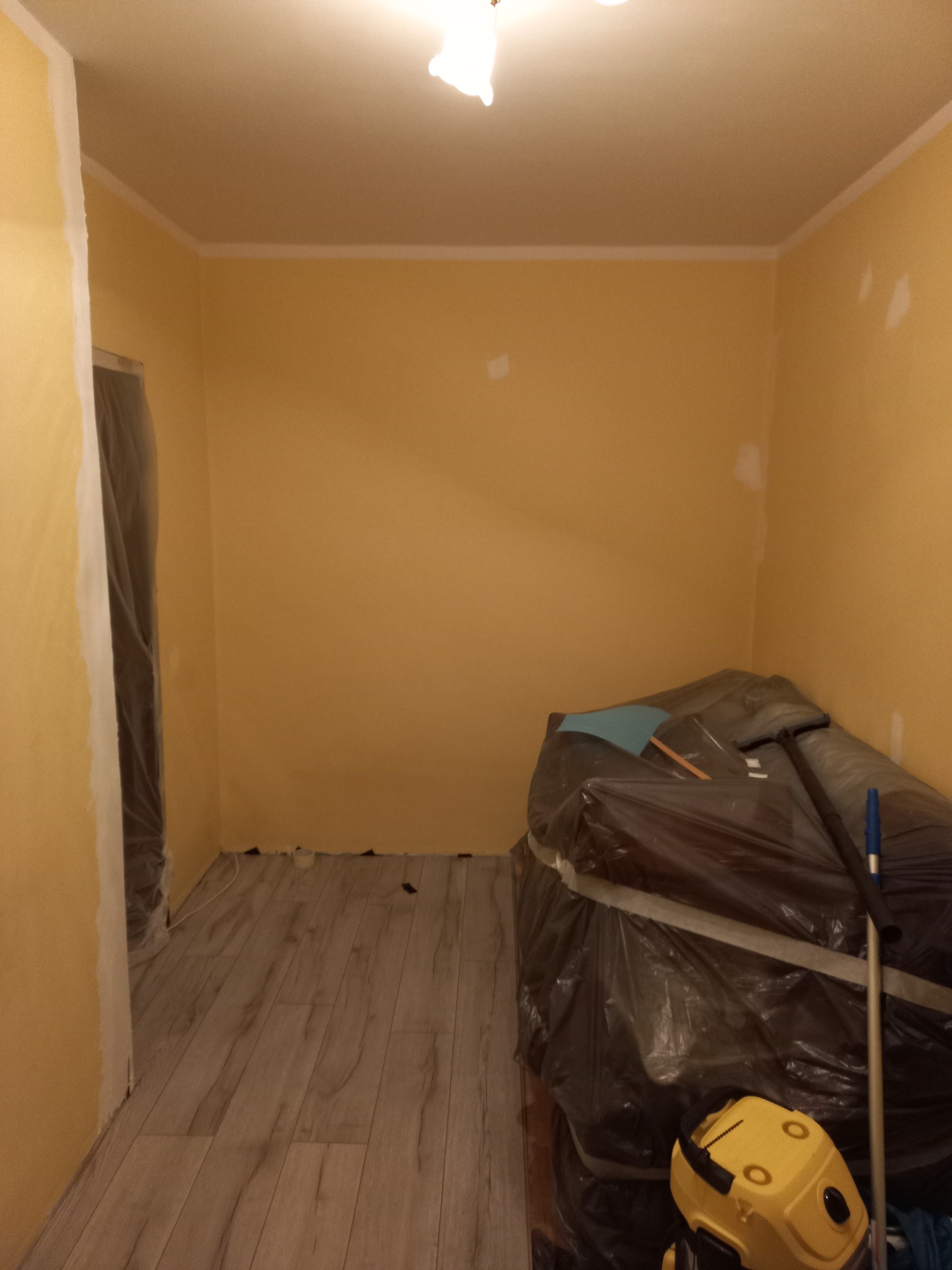 Pokój w trakcie malowania na żółto, z zabezpieczonymi folią meblami i widocznym odkurzaczem. Świeżo położone panele podłogowe i pomalowane listwy przypodłogowe.
