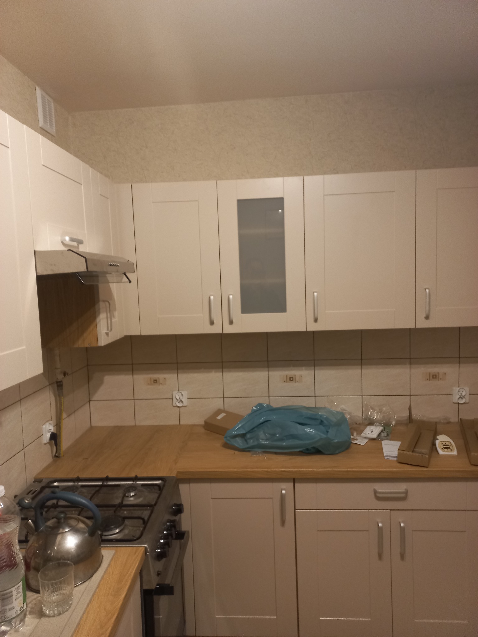 Jasna kuchnia z beżowymi szafkami i drewnianym blatem. Na ścianie płytki i tapeta. Widoczne akcesoria kuchenne i elementy montażowe. Ujęcie wnętrza po częściowym remoncie.