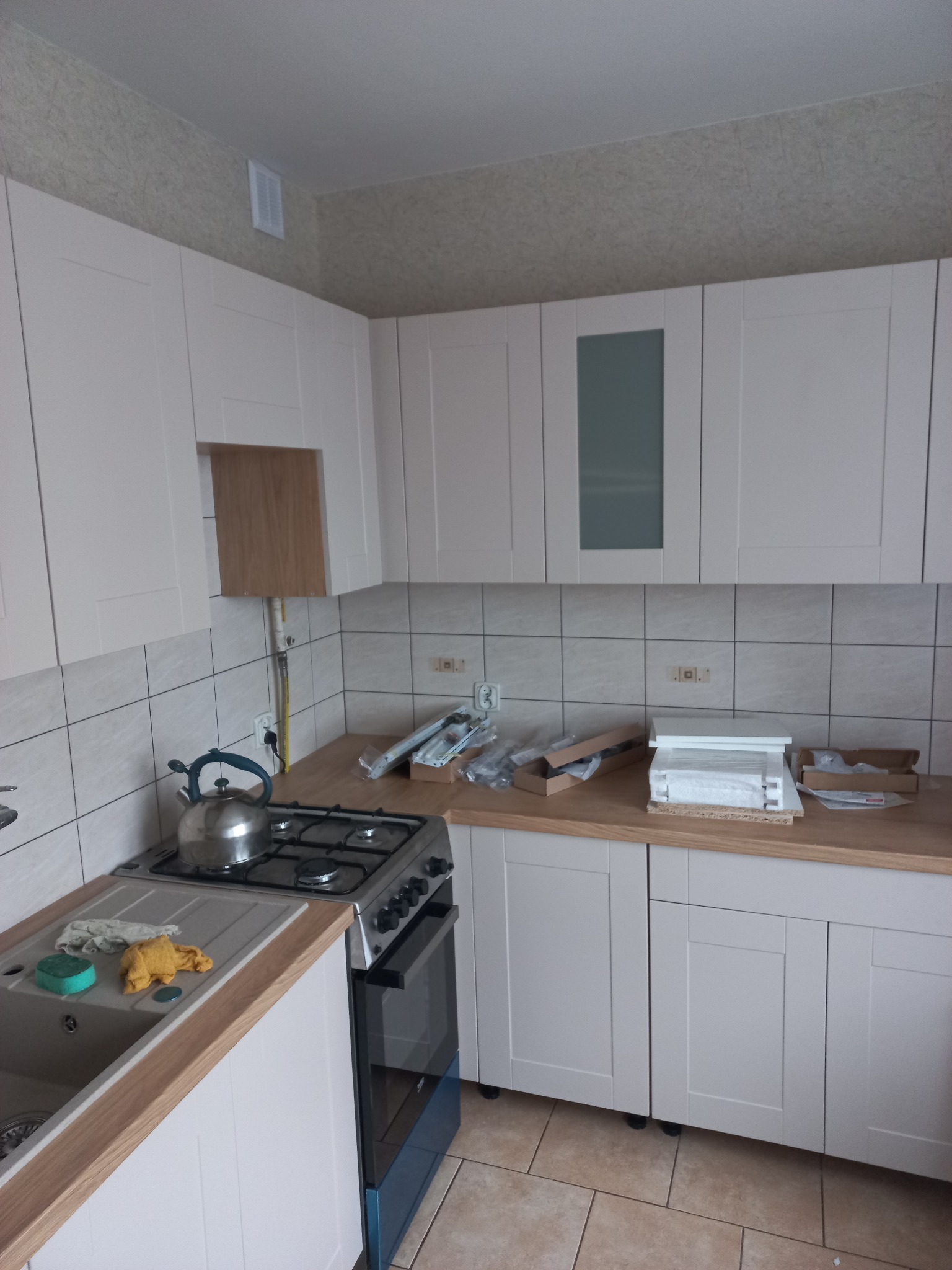 Jasna kuchnia z białymi szafkami i drewnianym blatem. Na blacie widoczne akcesoria montażowe. Na ścianie beżowe płytki i tapeta. Ujęcie z perspektywy.