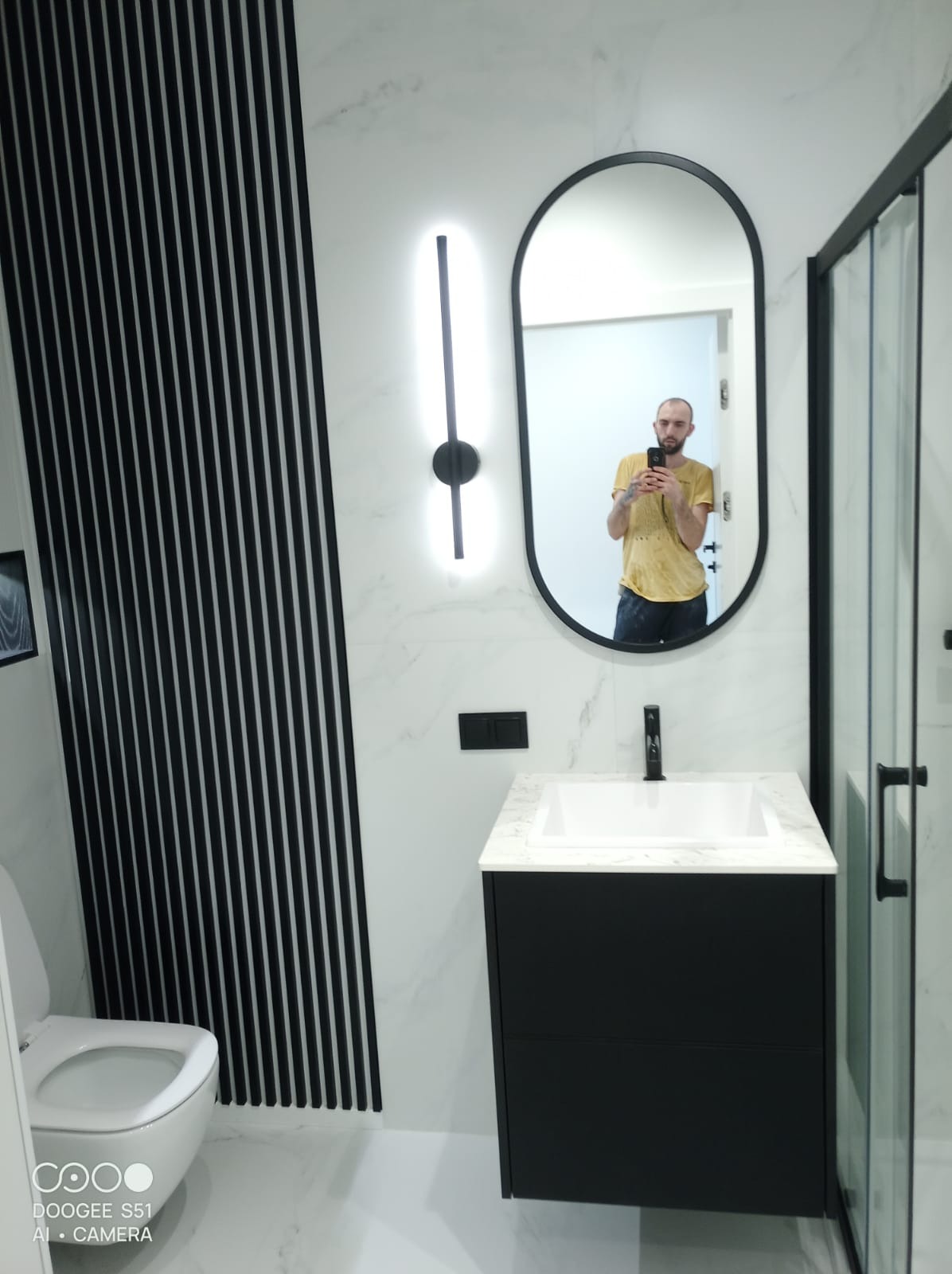 Nowoczesna łazienka z czarną armaturą i umywalką na szafce, marmurowe płytki, dekoracyjne listwy ścienne i podświetlane lustro owalne.