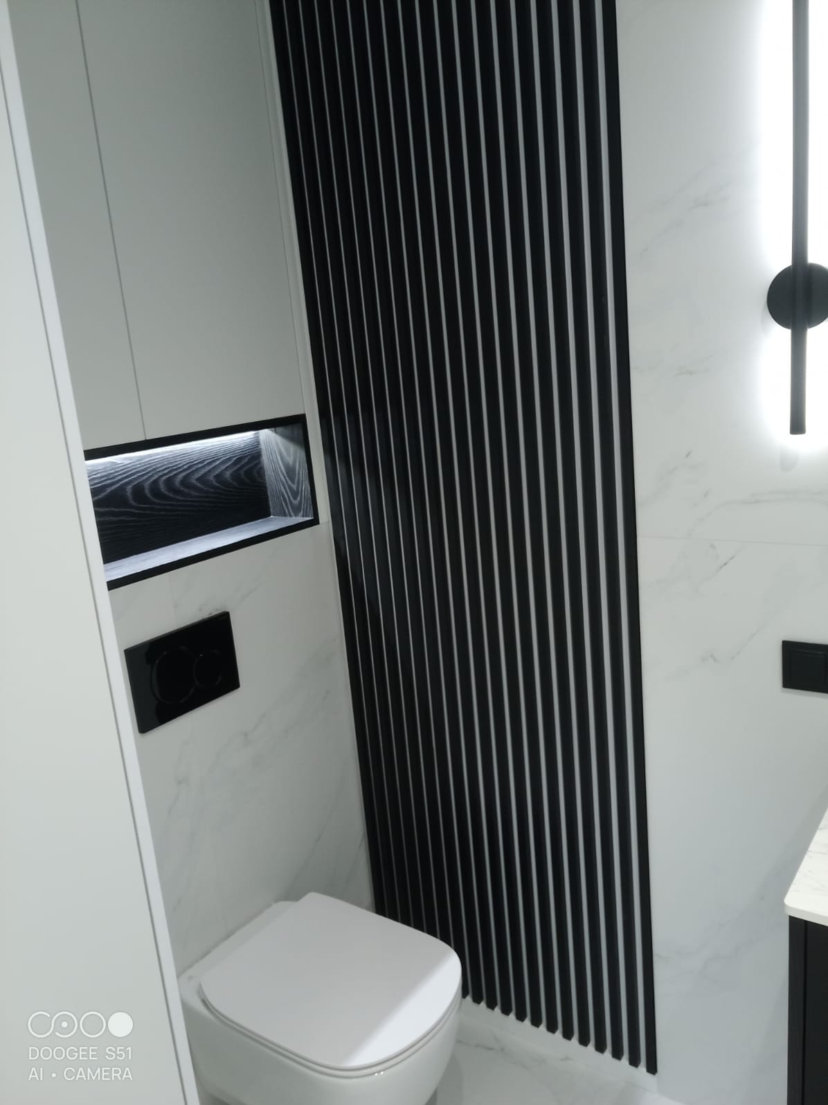 Nowoczesna łazienka po remoncie w Warszawie: białe marmurowe ściany, czarne pionowe lamele, podświetlana wnęka z drewnianym wykończeniem i podwieszana toaleta.