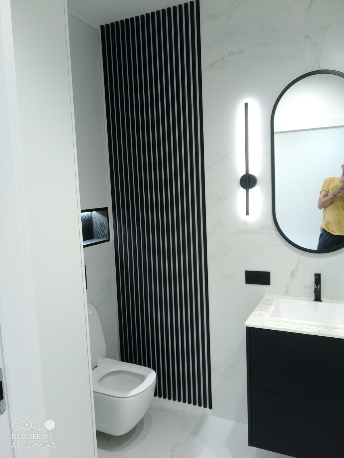 Nowoczesna łazienka z marmurowymi płytkami, czarnymi lamelami, owalnym lustrem z podświetleniem i czarną szafką umywalkową. Widoczny fragment toalety.