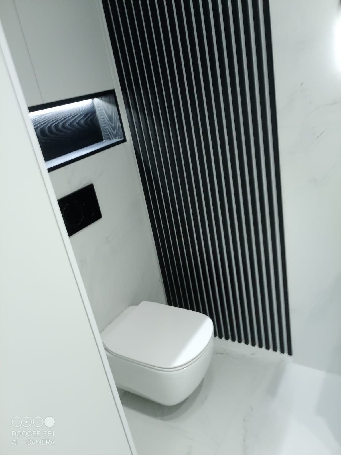 Nowoczesna, biała toaleta podwieszana w łazience z marmurowym wykończeniem i czarnymi, pionowymi lamelami dekoracyjnymi na ścianie.