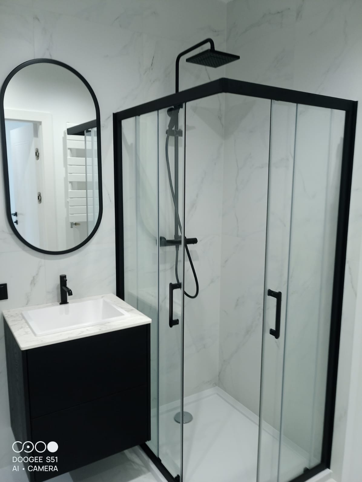 Nowoczesna łazienka z czarną armaturą: kabina prysznicowa z czarną ramą, umywalka z szafką i owalne lustro na tle marmurowych płytek. Minimalistyczny design.