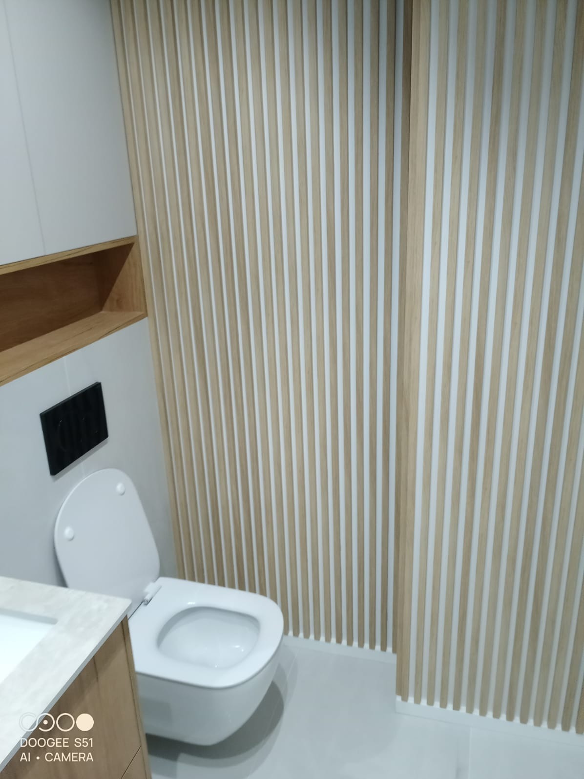 Nowoczesna łazienka z toaletą podwieszaną i dekoracyjnymi panelami ściennymi z drewna. Widoczna półka z drewna i białe płytki podłogowe.