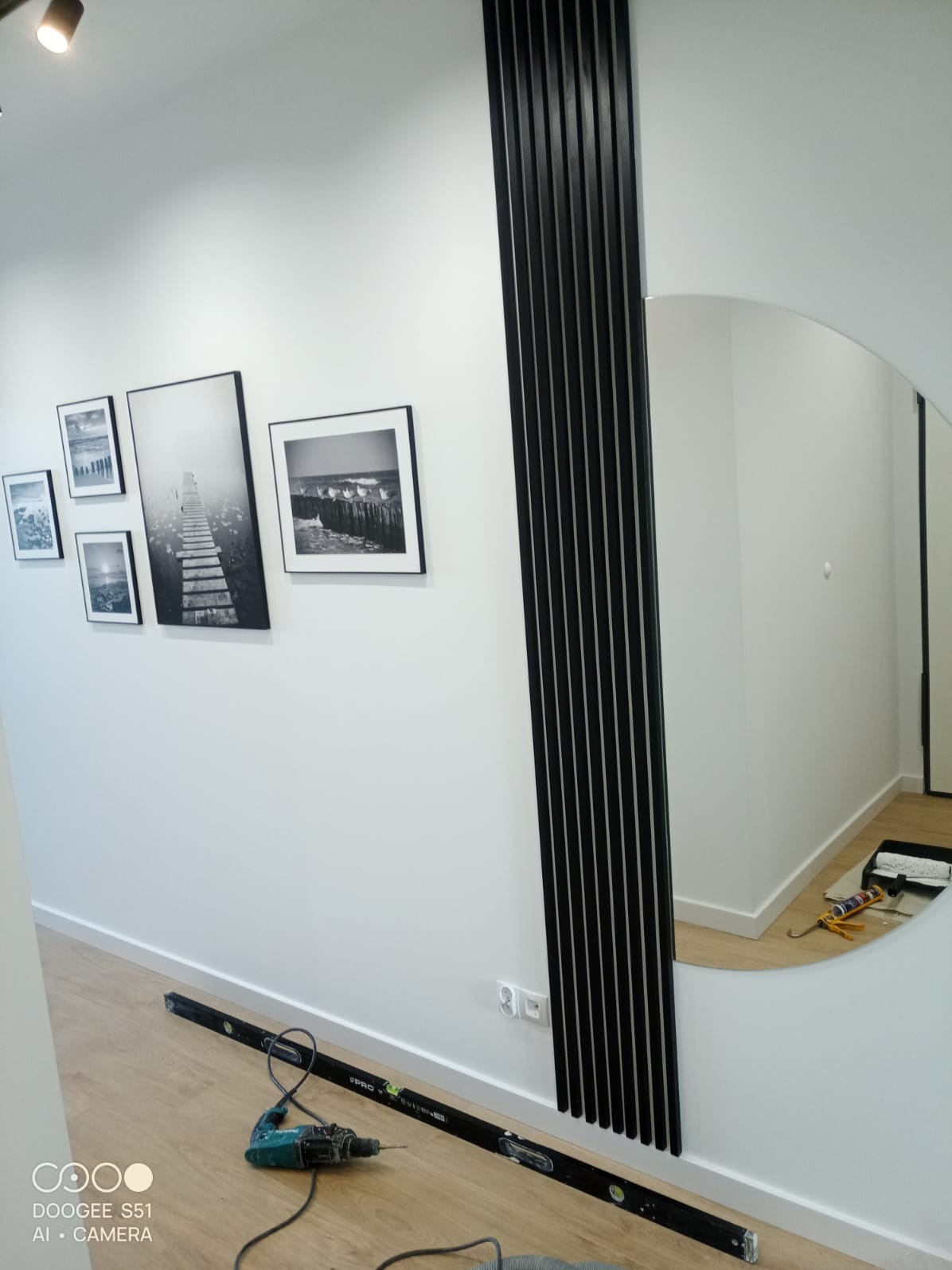 Nowoczesny salon z dekoracyjną ścianą z czarnych listew, lustrem i galerią czarno-białych zdjęć. Widoczne narzędzia wskazują na prace wykończeniowe.