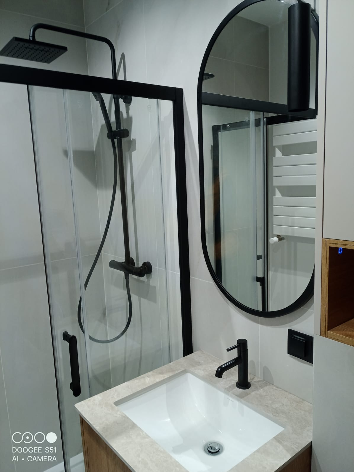 Nowoczesna łazienka z kabiną prysznicową typu walk-in, owalnym lustrem w czarnej ramie i umywalką na blacie z drewnianą szafką. Armatura w kolorze czarnym.