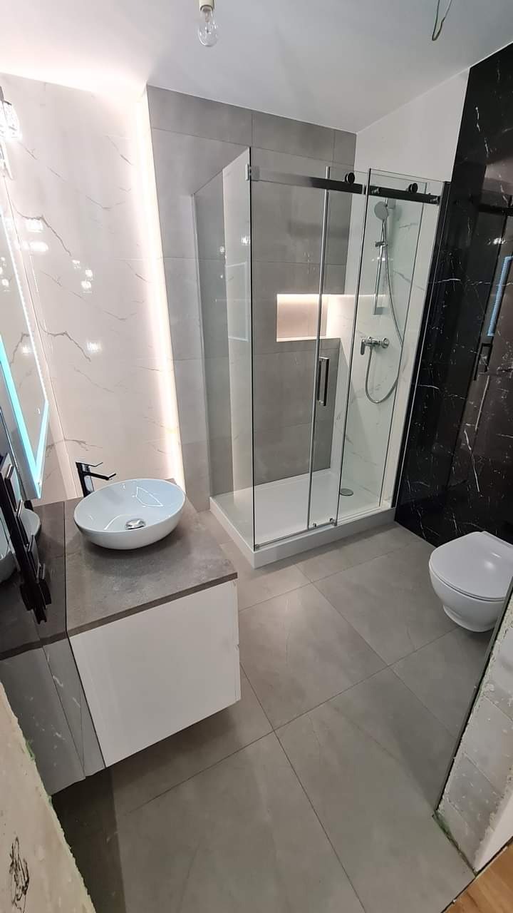 Nowoczesna łazienka z kabiną prysznicową typu walk-in, umywalką nablatową i podświetleniem LED. Czarno-białe marmurowe płytki i szara podłoga tworzą elegancki kontrast.