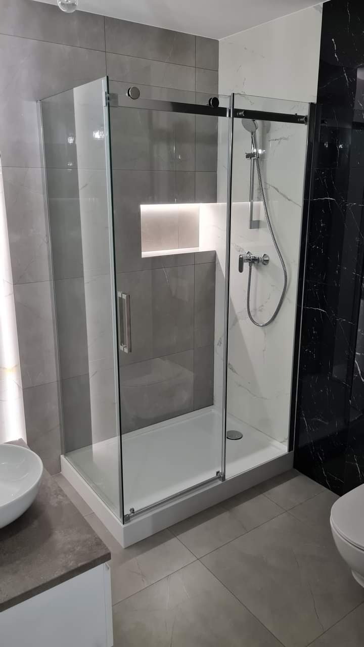 Nowoczesna kabina prysznicowa z przesuwnymi drzwiami, wykończona szarymi i białymi płytkami z marmurowym wzorem. W ścianie wnęka z oświetleniem LED.