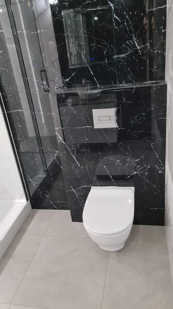 Elegancka łazienka z czarną marmurową ścianą, podwieszaną toaletą i szklaną kabiną prysznicową. Minimalistyczny design, jasne płytki podłogowe.