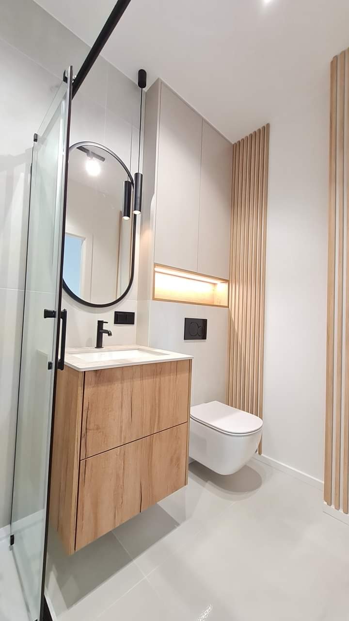 Nowoczesna łazienka z drewnianą szafką pod umywalką, owalnym lustrem z czarną ramą i podwieszaną toaletą. Ściana zdobiona drewnianymi lamelami.
