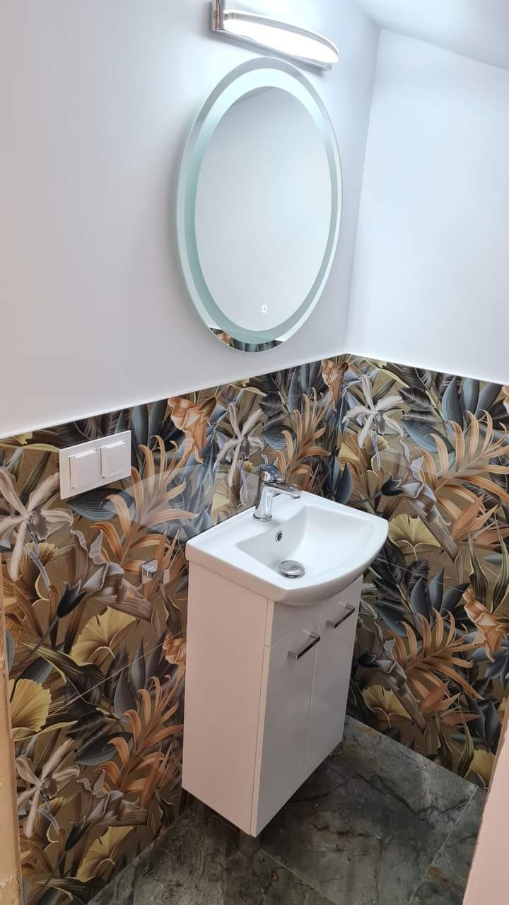 Biała szafka z umywalką, okrągłe lustro z podświetleniem i wzorzyste płytki z motywem roślinnym w wyremontowanej łazience. Nowoczesny wystrój.