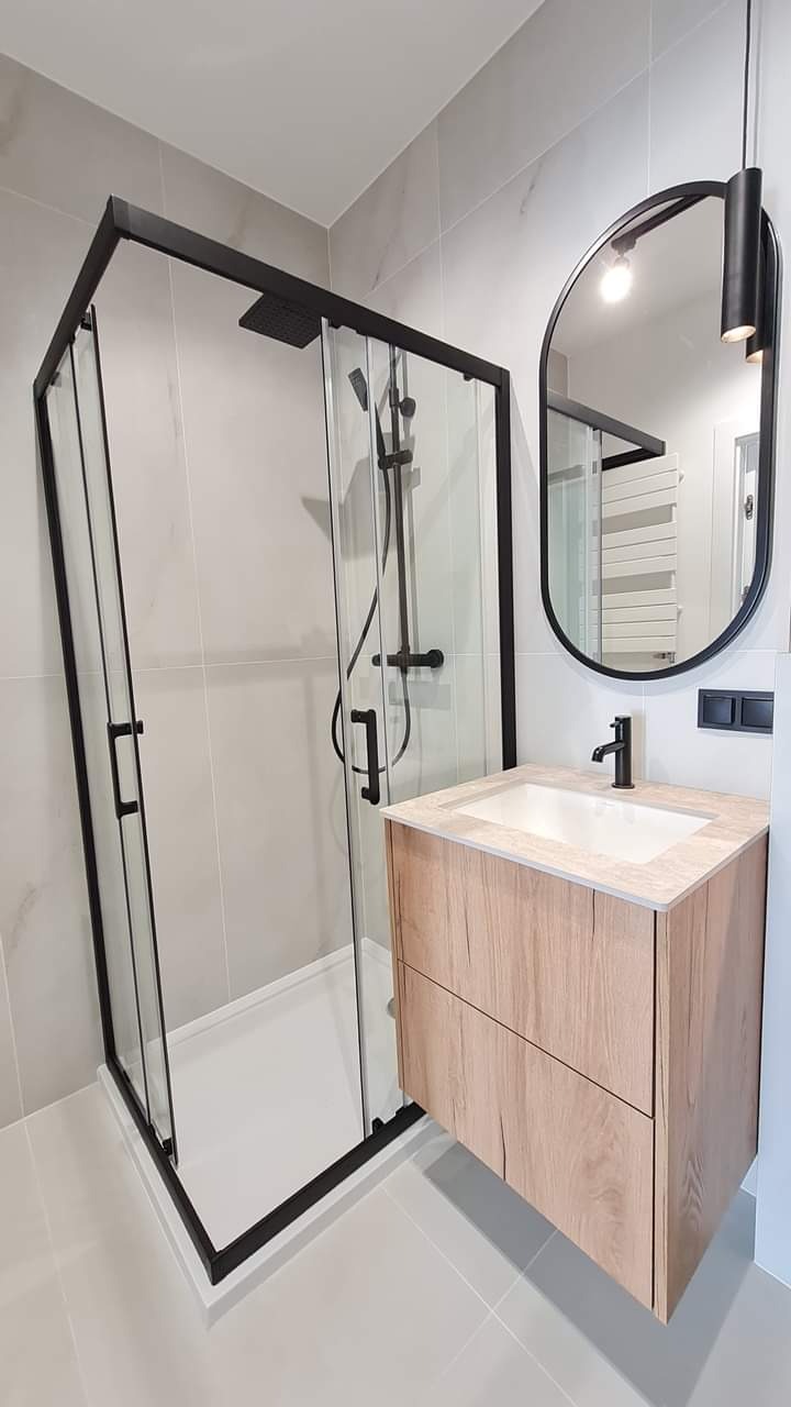 Nowoczesna łazienka z kabiną prysznicową typu walk-in z czarnymi profilami, umywalką z szafką z drewnopodobnym frontem i owalnym lustrem z czarną ramą.