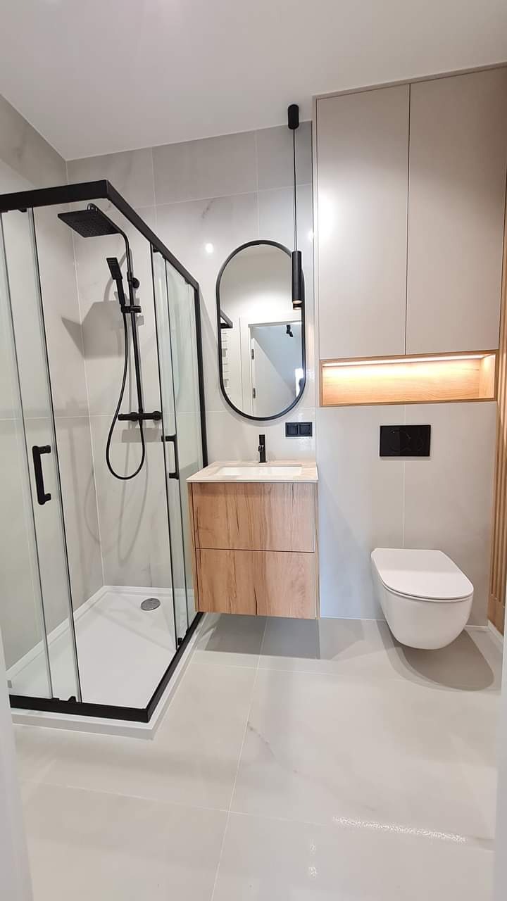 Nowoczesna łazienka z kabiną prysznicową w czarnej ramie, owalnym lustrem i drewnianą szafką pod umywalką. Minimalistyczny design z elementami oświetlenia.