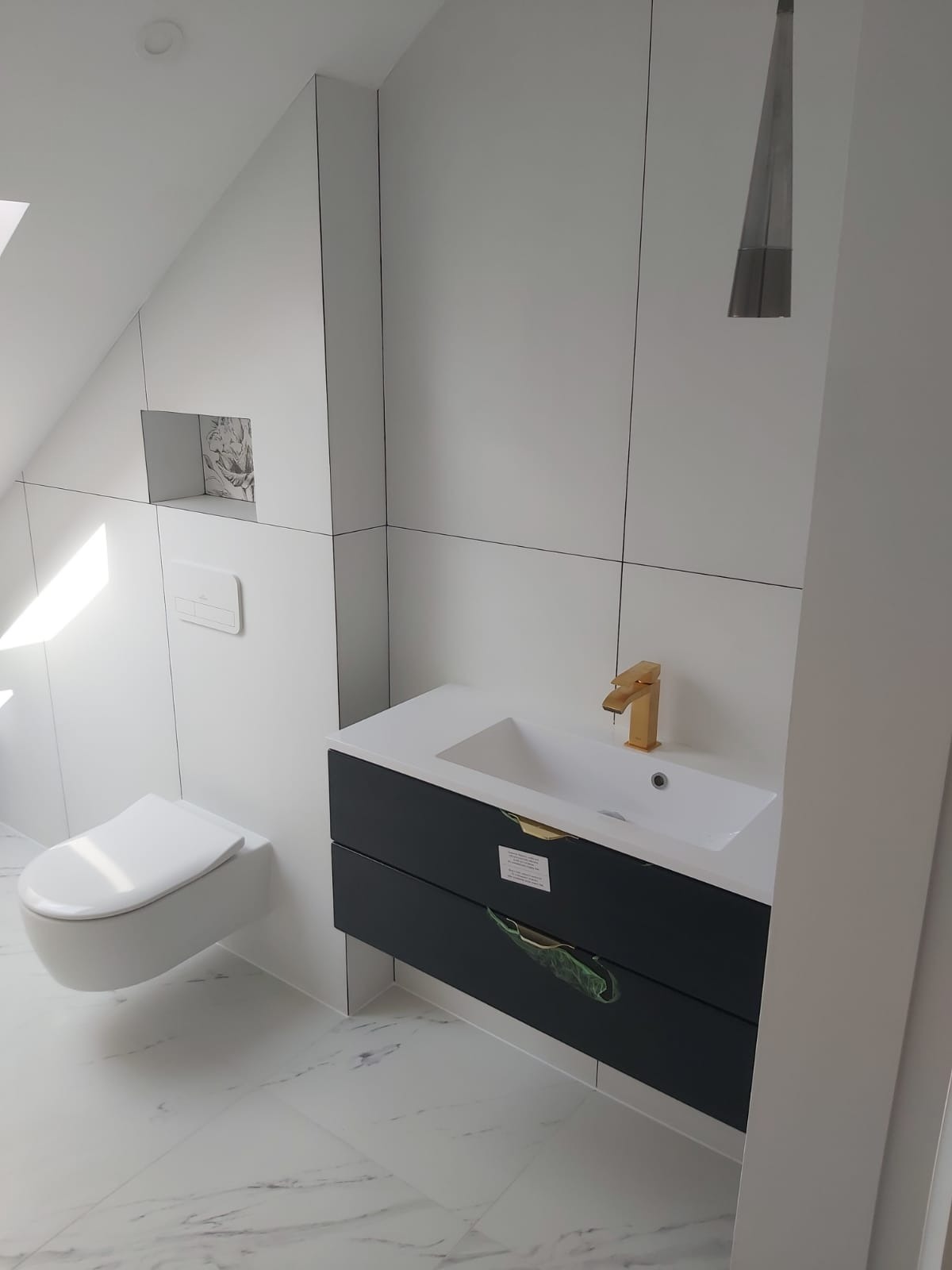 Nowoczesna łazienka na poddaszu z białą armaturą, czarną szafką pod umywalką i złotą baterią. Minimalistyczny design, marmurowe płytki podłogowe.