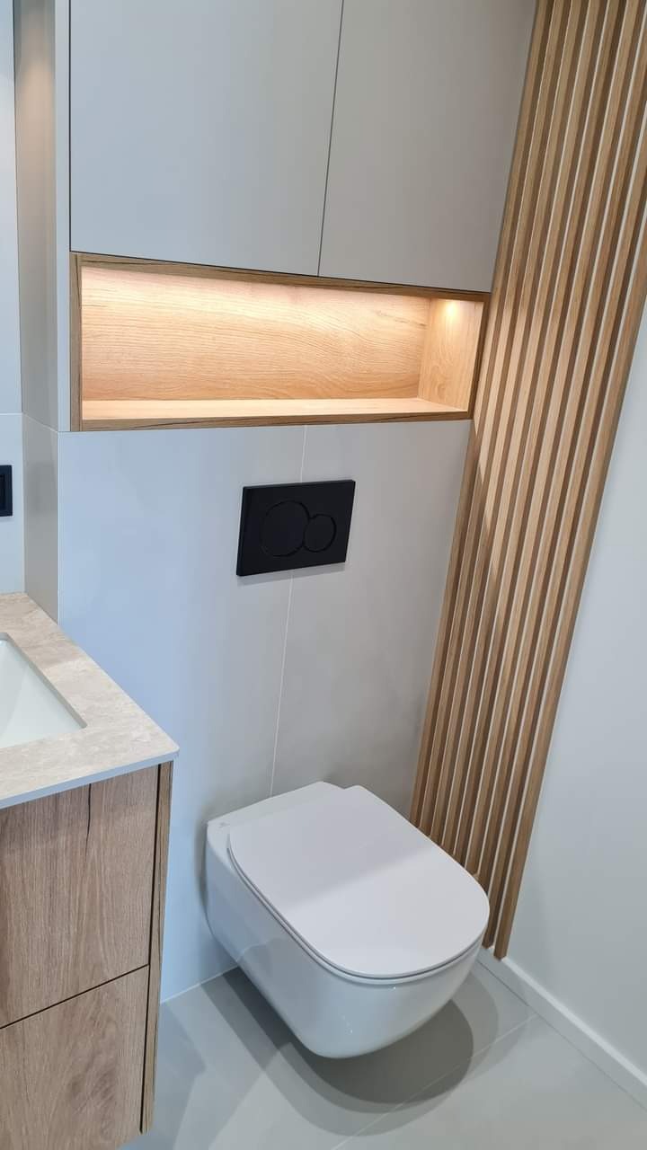 Nowoczesna łazienka z podwieszaną toaletą, drewnianymi lamelami i szafkami z oświetleniem LED. Minimalistyczny design z akcentami drewna.