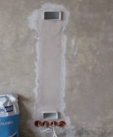 Bruzda w ścianie po instalacji elektrycznej, z widocznymi puszkami i przewodami, przygotowana do szpachlowania. Obok worek gipsu.
