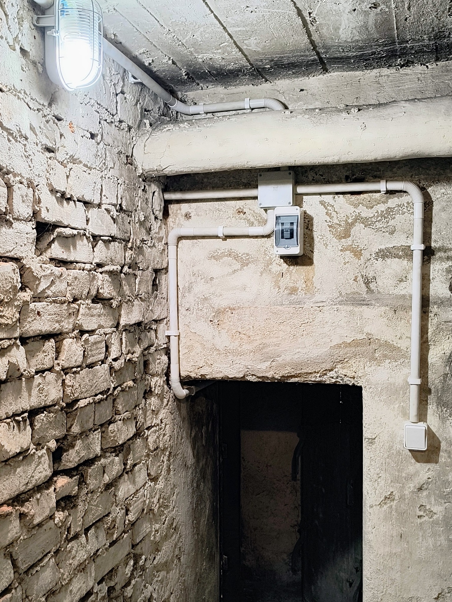 Białe rury i skrzynka elektryczna na ścianie piwnicy z cegły, oświetlone lampą. Instalacja w surowym wnętrzu, widok na ciemne wejście.